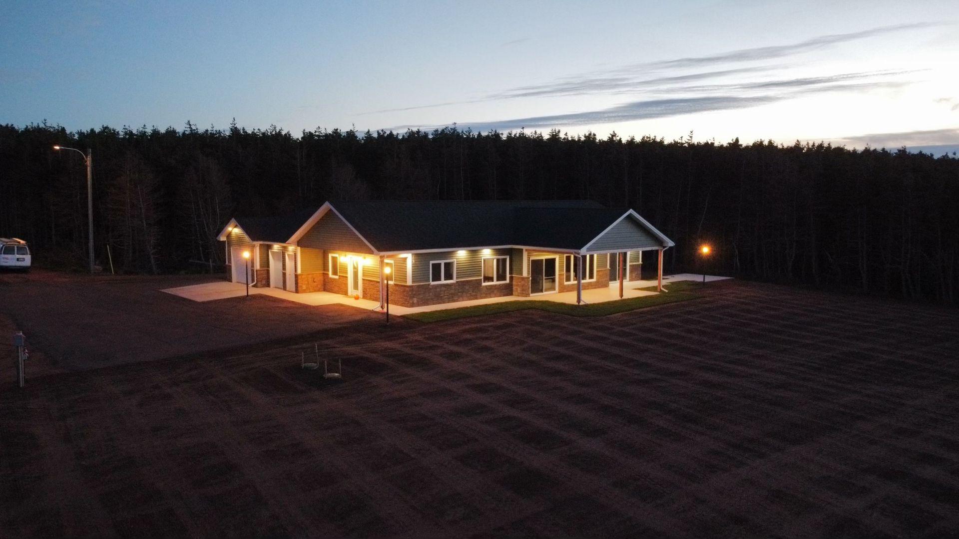 Single-story house with exterior lights on, surrounded by trees at dusk.