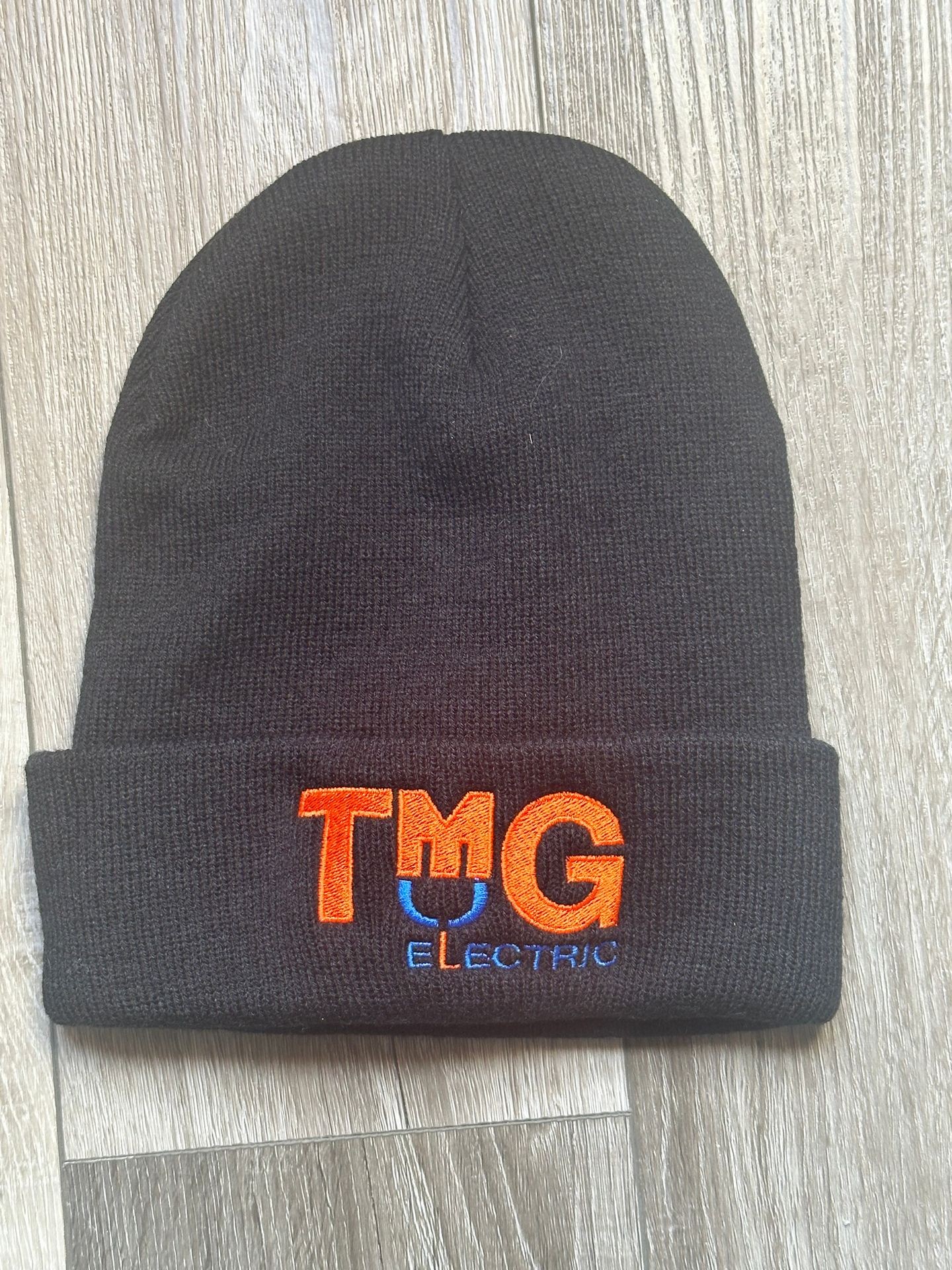 Black beanie with 'TMG Electric' embroidered in orange and blue on a wooden surface.