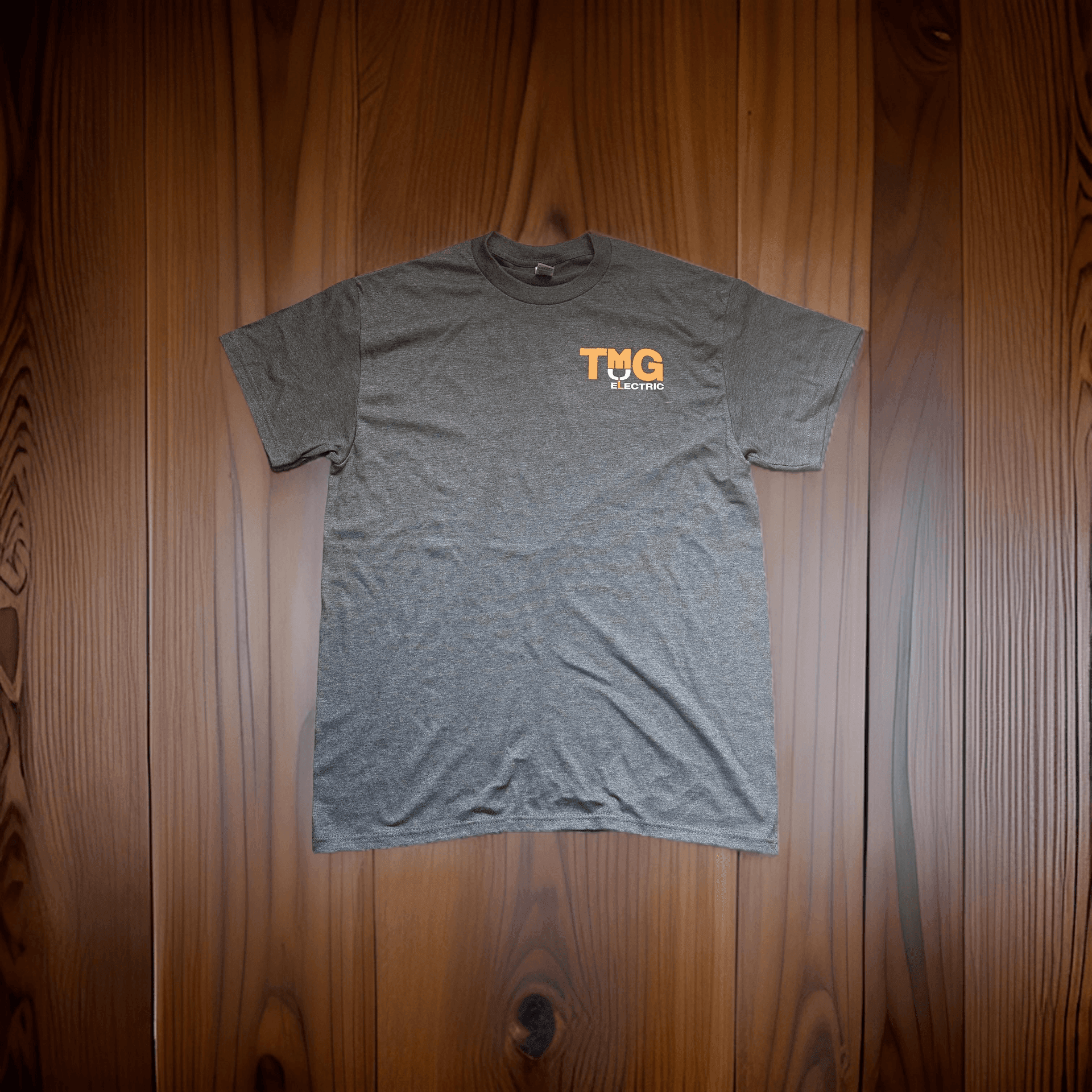 Gray T-shirt with orange 'TMG Electric' logo on wooden background.